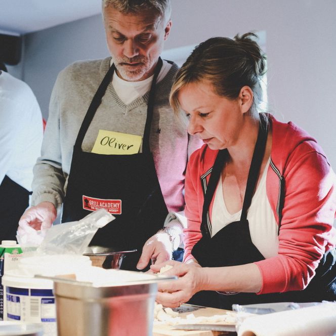 Eine Frau schneidet Fetakäse klein, daneben stehen zwei Männer an einer Arbeitsplatte.