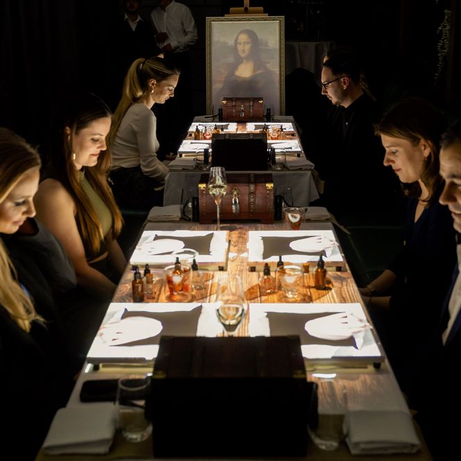 Sechs Personen sitzen an einem langen, beleuchteten Tisch mit Schachbrettern, im Hintergrund ein Bild der Mona Lisa